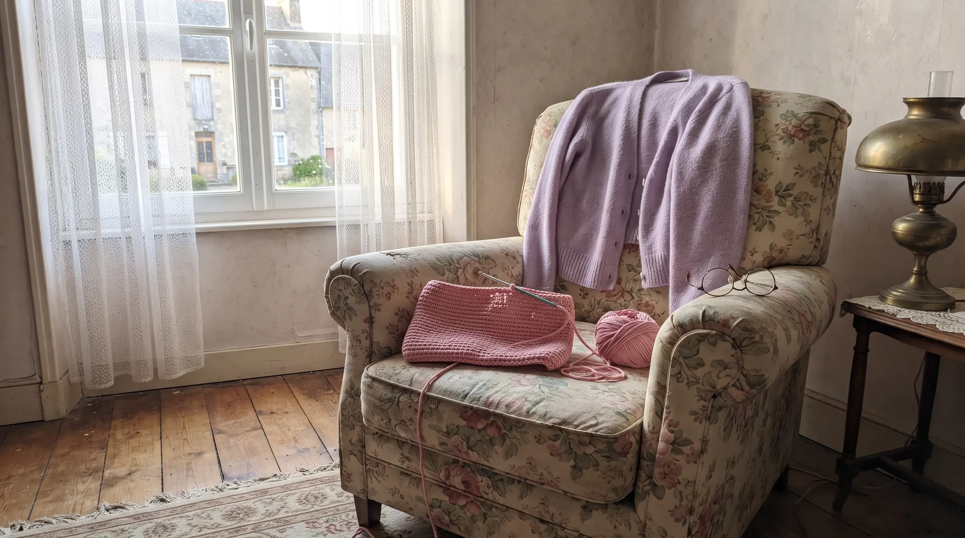 Un fauteuil à fleurs vide près de la fenêtre, un ouvrage au crochet rose posé sur l'assise, un gilet lavande sur le dossier
